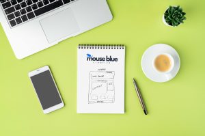 Small business website design strategy sketch on notepad with laptop, phone, and coffee on desk.